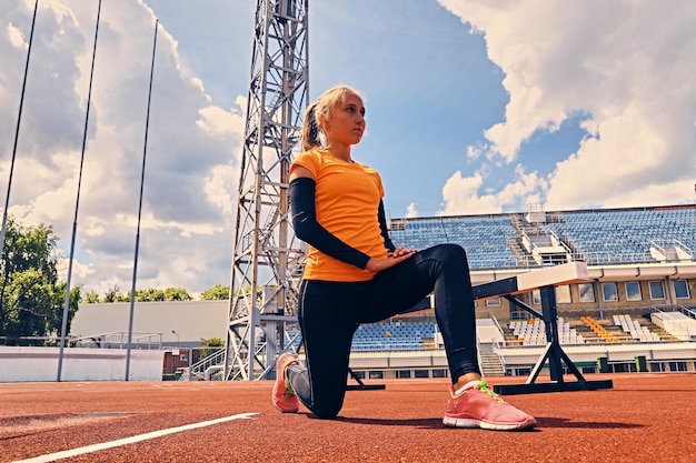 Blonde sportieve vrouwelijke loper naar snelle startpositie op een stadion.