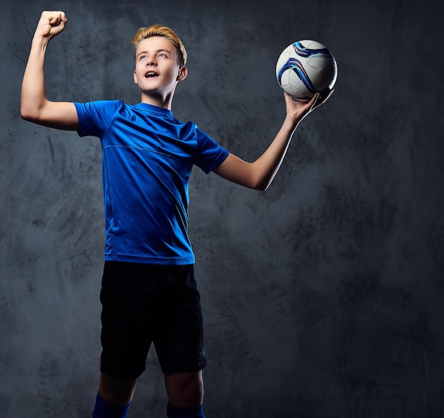 Blonde tiener, voetballer gekleed in een blauw uniform houdt een bal vast.