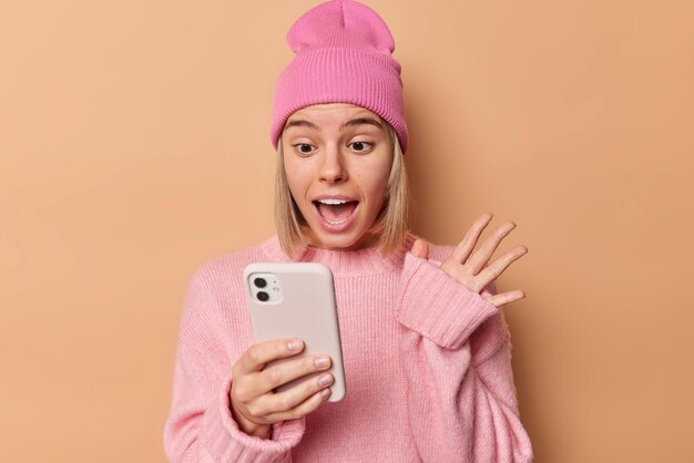 Blonde verraste vrouw ziet er opgewonden uit en reageert op geweldige nieuwsrollen sociale media pagina's via smartphone houdt handpalm omhoog draagt vrijetijdskleding geïsoleerd over beige achtergrond Wow dat is geweldig