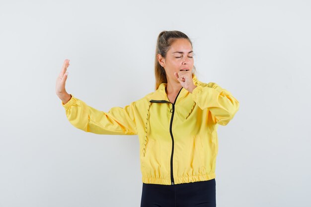 Blonde vrouw die doet alsof ze in een geel bomberjack en een zwarte broek zingt en geamuseerd kijkt