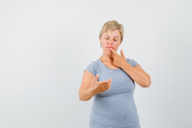 Blonde vrouw die doet alsof ze naar de telefoon kijkt en haar vinger aan de lippen zet in een lichtblauw t-shirt en peinzend, vooraanzicht kijkt.