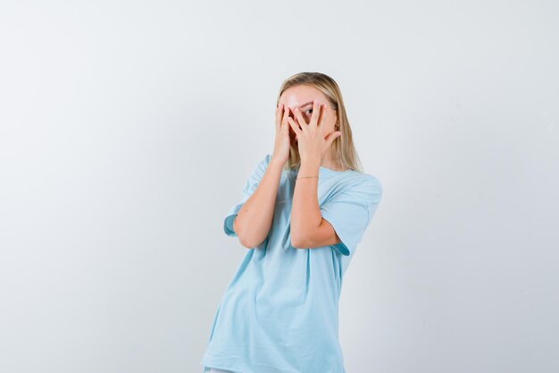 Blonde vrouw die door vingers in blauw t-shirt kijkt en er schattig uitziet