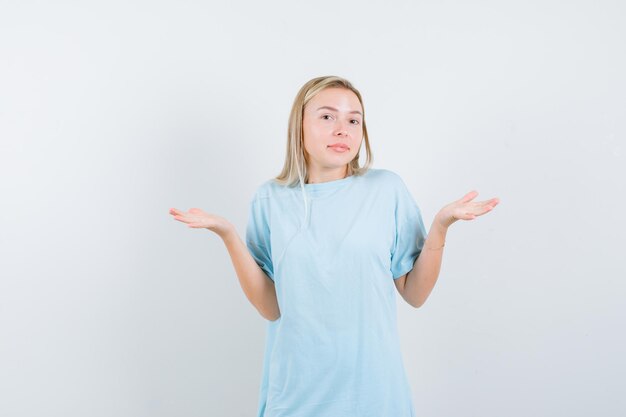 Blonde vrouw die hulpeloos gebaar in blauw t-shirt toont en verward kijkt