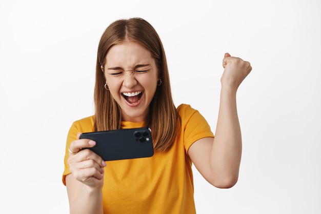 Blonde vrouw die smartphone horizontaal houdt, mobiel videospel speelt en vuistpomp schreeuwt van vreugde en succes, overwinning viert, witte muur