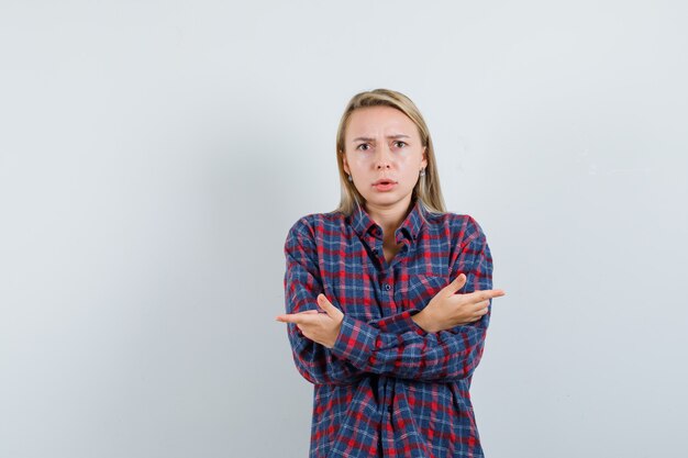 Blonde vrouw die verschillende richtingen wijst met wijsvingers in geruit overhemd en verbaasd, vooraanzicht kijkt.
