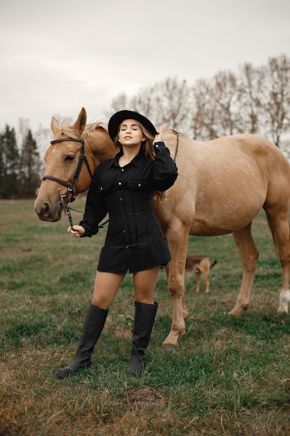 Blonde vrouw en bruin paard staan in het veld. Vrouw die zwarte kleren en hoed draagt. Vrouw die het paard aanraakt.