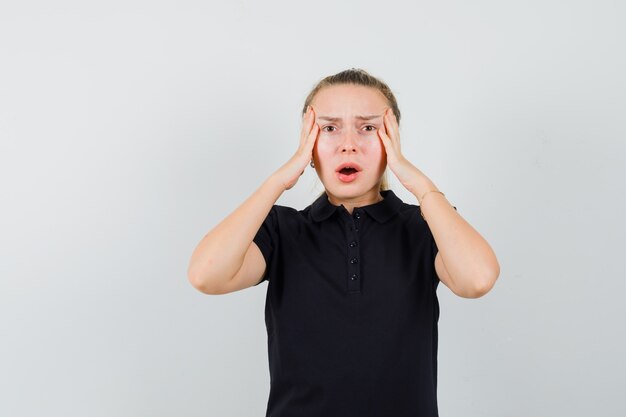 Blonde vrouw houdt haar handen op haar hoofd met open mond in zwart t-shirt en kijkt geïrriteerd