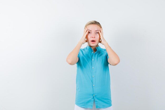 Blonde vrouw houdt handen op het hoofd in blauwe blouse en kijkt verbijsterd geïsoleerd