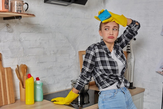 Gratis foto blonde vrouw in beschermende handschoenen met vodden die elektrisch fornuis thuis keuken schoonmaken. meisje dat zwart glanzend oppervlak van keukenblad wast. concept van huishoudelijk werk.