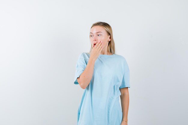 Blonde vrouw in blauw t-shirt die de mond bedekt met de hand, wegkijkend en verbaasd kijkend