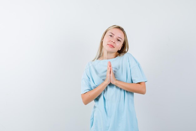 Blonde vrouw in blauw t-shirt met namaste gebaar en ziet er schattig uit