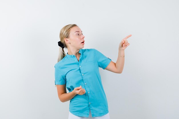 Blonde vrouw in blauwe blouse naar rechts wijzend en verbaasd geïsoleerd kijken