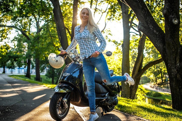 Blonde vrouw in casual kleding bezit op moto scooter over groene bomen achtergrond.