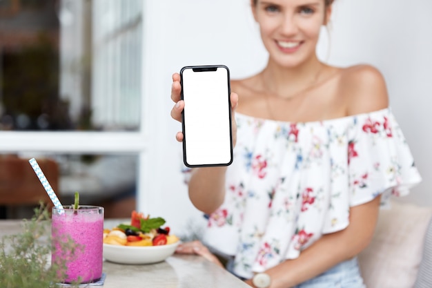 Blonde vrouw in gebloemde jurk in café met het lege smartphonescherm
