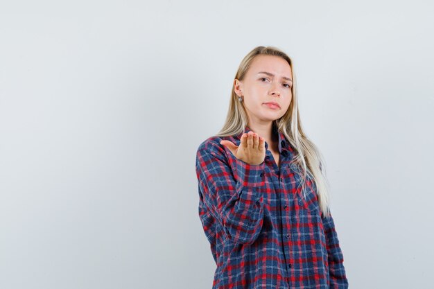 Blonde vrouw in geruit overhemd die één hand naar camera uitrekt als proberend om iets te ontvangen en nieuwsgierig, vooraanzicht kijkt.