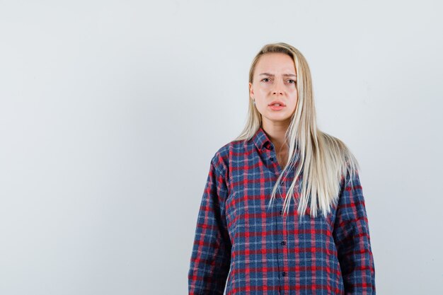 Blonde vrouw in geruit overhemd die zich rechtop en grimassen bevindt en ontevreden, vooraanzicht kijkt.