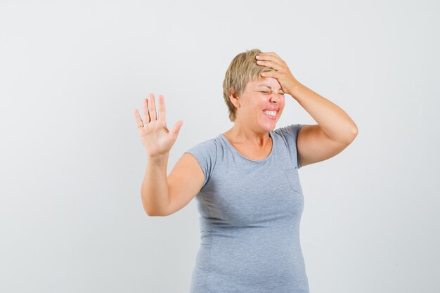 Blonde vrouw in lichtblauw t-shirt met weigering gebaar en houdt haar hand op het hoofd en kijkt geïrriteerd, vooraanzicht.