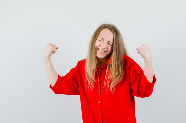 Blonde vrouw in rood shirt winnaar gebaar tonen en op zoek gelukkig,