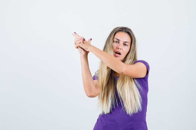 blonde vrouw in violet t-shirt pistool gebaar tonen en op zoek zelfverzekerd, vooraanzicht.