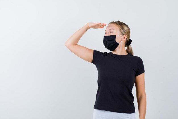 Blonde vrouw in zwart t-shirt, witte broek, zwart masker kijkt ver weg