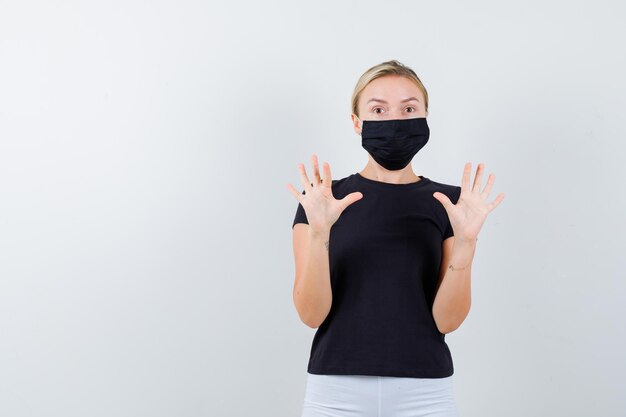 Blonde vrouw in zwart t-shirt, witte broek, zwart masker met opgeheven handpalmen