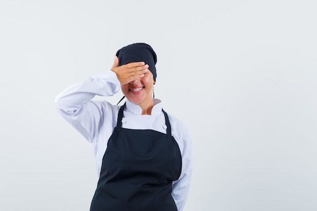 Blonde vrouw in zwarte kok uniform bedekkend oog met handen en verlegen kijkt