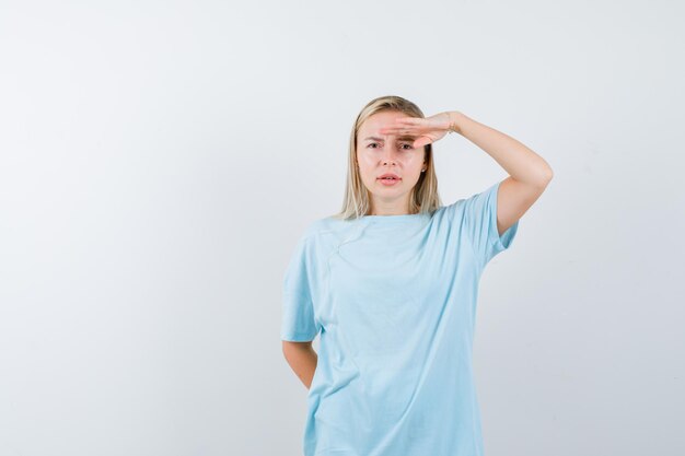 Blonde vrouw kijkt ver weg met de hand boven het hoofd in blauw t-shirt en kijkt gefocust