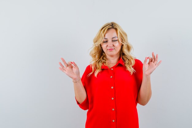 Blonde vrouw mediteren terwijl ze ogen in rode blouse sluit en er ontspannen uitziet.