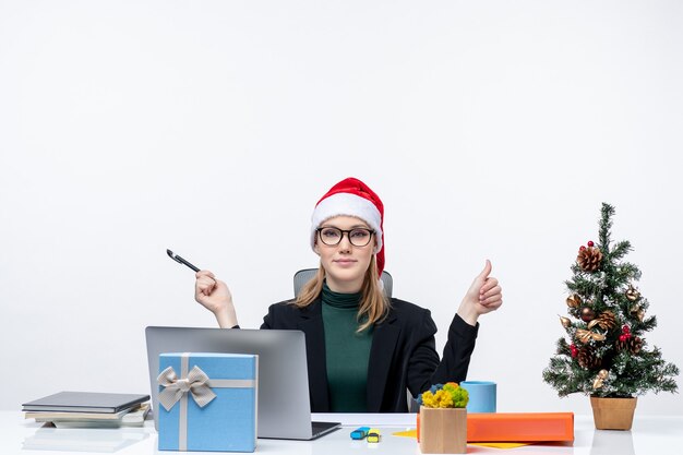 Blonde vrouw met een kerstman hoed zittend aan een tafel met een kerstboom en een cadeau