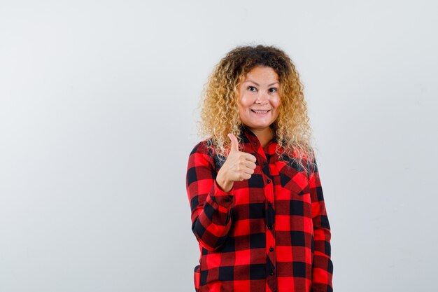 Blonde vrouw met krullend haar duim opdagen in geruit overhemd en joviaal kijken. vooraanzicht.