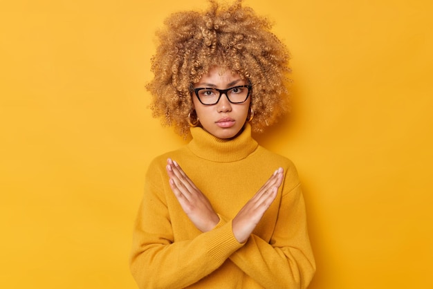 Blonde vrouw met krullend haar toont kruis maakt stopgebaar zegt nee met boze uitdrukking weigert iets draagt een bril en trui met lange mouwen geïsoleerd op gele achtergrond. Het is taboe.