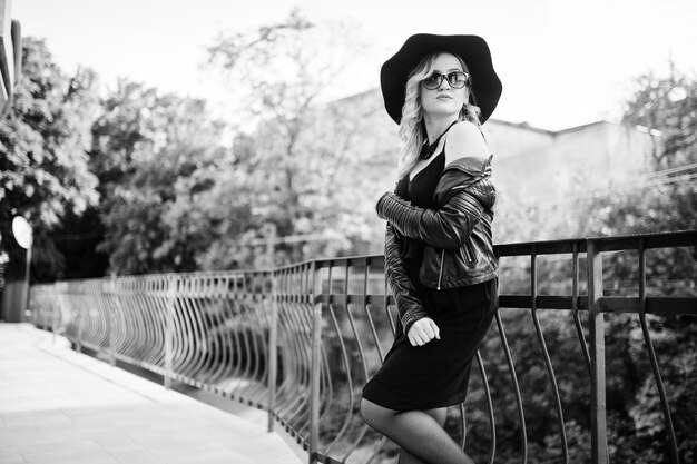 Blonde vrouw op zwarte jurk leren jas zonnebril kettingen en hoed tegen balustrades