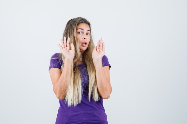 blonde vrouw palmen in overgave gebaar in violet t-shirt tonen en bang op zoek. vooraanzicht.