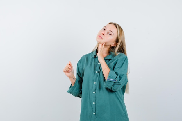 Blonde vrouw poseert terwijl ze haar kin aanraakt in een groen shirt en er charmant uitziet