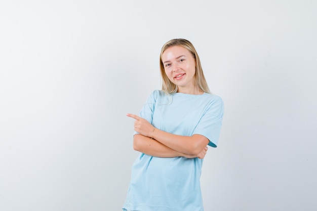 Blonde vrouw staande armen gekruist, naar links wijzend met wijsvinger in blauw t-shirt