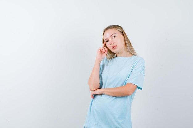 Blonde vrouw staat in denkende houding, wijsvinger op de tempel in blauw t-shirt