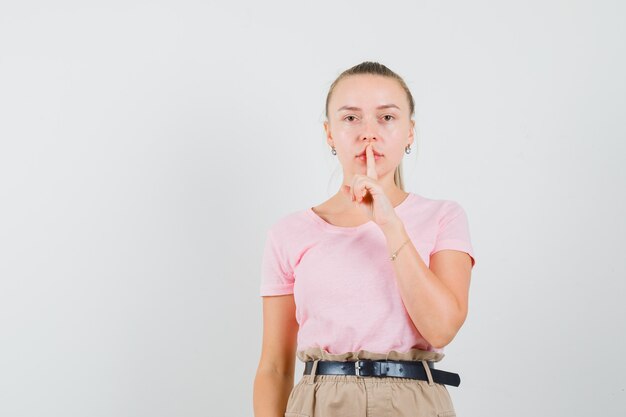 Blonde vrouw stilte gebaar in t-shirt, broek tonen en voorzichtig kijken. vooraanzicht.