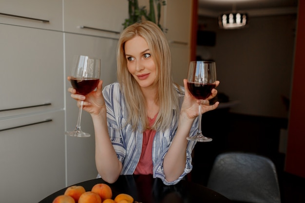 Blonde vrouw tases twee glazen rode wijn in haar eigen keuken.
