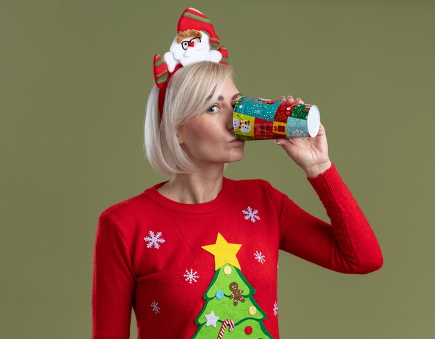 Blonde vrouw van middelbare leeftijd dragen hoofdband van de kerstman en kerst trui kijken camera koffie drinken uit plastic kerst koffiekopje geïsoleerd op olijfgroene achtergrond