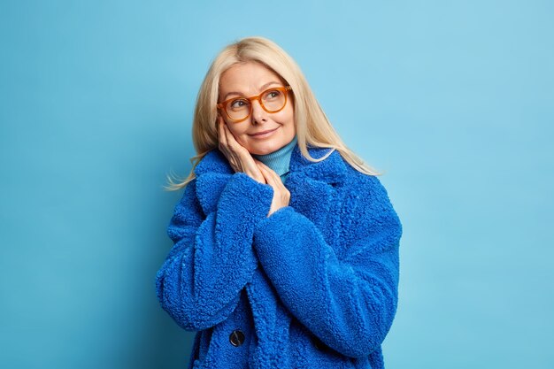 blonde vrouw van middelbare leeftijd in brillen herinnert zich iets aangenaams houdt de handen in de buurt van het gezicht denkt na over de toekomst kijkt opzij glimlacht draagt zachtjes warme blauwe winterjas.