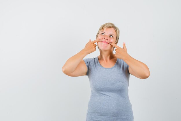 Blonde vrouw wijst naar haar lippen met haar wijsvingers en kijkt omhoog in lichtblauw t-shirt en ziet er vrolijk uit. vooraanzicht.