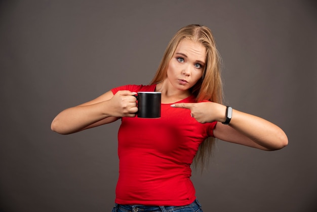 Gratis foto blonde vrouw wijzend op lege beker op zwarte muur.