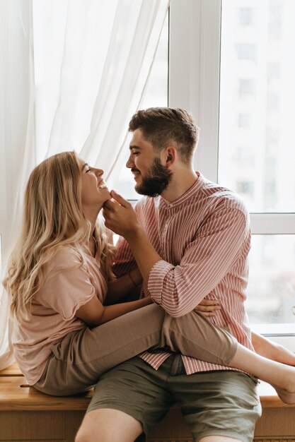 Blonde vrouw zit op haar vriendje en lacht. Man streelt met tederheid het gezicht van zijn geliefde. Portret van paar tegen venster.