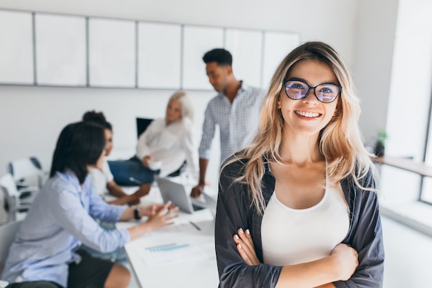 Gratis foto blonde vrouwelijke uitvoerende poseren met glimlach en armen gekruist tijdens brainstorm met managers. indoor portret van europese student tijd doorbrengen in hal met aziatische en afrikaanse vrienden.