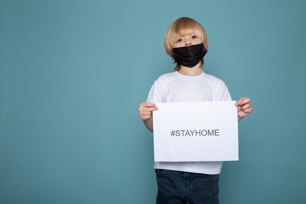 Blondejongen met zwarte beschermende het verblijfshashtag van de maskerholding tegen coronavirus op blauwe muur