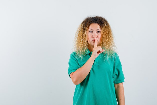 Blondevrouw met krullend haar die stiltegebaar in groen T-shirt tonen en zorgvuldig kijken, vooraanzicht.