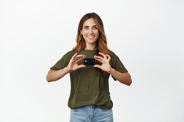 Boodschappen doen. Glimlachende vrouw van middelbare leeftijd die een creditcard met korting toont, er gelukkig uitziet, contactloos betalen adviseert, iets koopt, over een witte achtergrond staat.