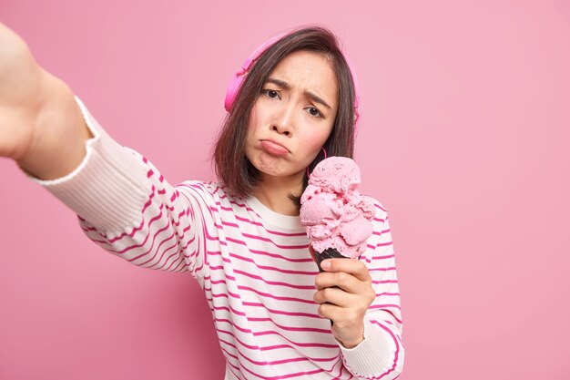 Boos Aziatische tienermeisje kantelt hoofd ziet er helaas maakt selfie portret houdt lekker ijs kantelt hoofd luistert muziek via draadloze koptelefoon gekleed in gestreepte trui geïsoleerd over roze muur