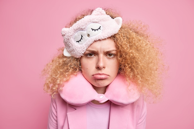 Boos beledigd mooie vrouw met krullend haar ziet er helaas haat vroeg ontwaken draagt slaapmasker reiskussen om nek heeft ellendige gezichtsuitdrukking geïsoleerd over roze muur