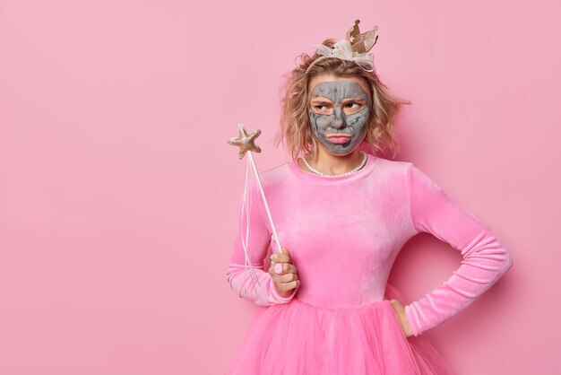 Boos beledigde jonge vrouw kijkt ongelukkig weg draagt kroon en feestelijke jurk past schoonheidsmasker toe voor teintverzorging houdt toverstaf poseert tegen roze achtergrond kopieer ruimte voor je promo